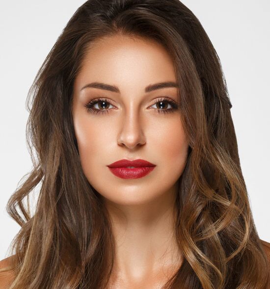 Confident woman with long wavy brown hair, bold red lipstick, and flawless skin looking directly at the camera.