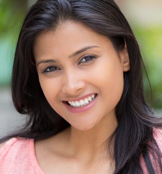 miling woman with long dark hair wearing a light pink top, posing outdoors with a natural, radiant look.