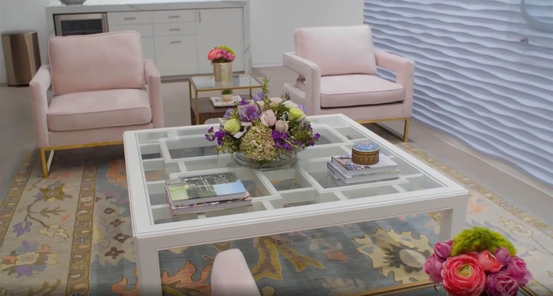 Modern waiting area with pastel pink chairs, a glass coffee table decorated with flowers and books, and a patterned rug creating a welcoming atmosphere.